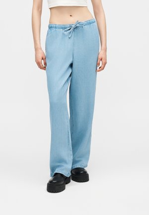 Γυναίκα που φοράει ανοιχτό μπλε τζιν παντελόνια με φαρδιά μπατζάκια, ελαστική μέση και κορδόνια, σε συνδυασμό με μαύρα παπούτσια με χοντρή σόλα.