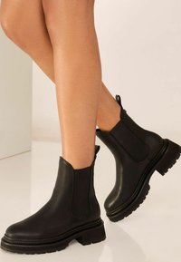 Fekete bokacsizmák sima bőrből, rugalmas oldalsó panelekkel, vastag talppal, húzófüllel és mintázott talppal a tapadás érdekében.