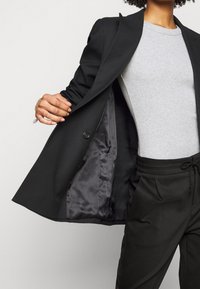 Svart skräddarsydd blazer med satininteriör, visad öppen, över en ljusgrå skjorta och svarta byxor med elastiskt midjeband.