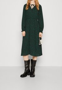 Grön blommig klänning med långa ärmar, V-ringning och knytbrosch, kombinerad med svarta knähöga stövlar med klumpig sula.