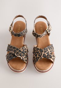 Paire de sandales pour femmes avec brides croisées imprimé léopard et brides de cheville avec boucles dorées sur fond beige.