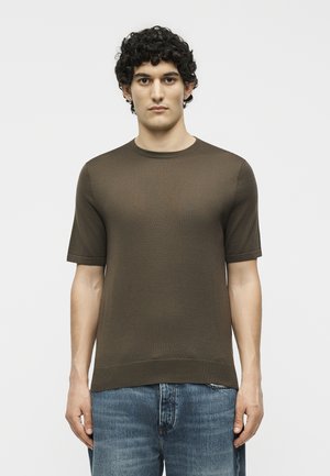 Νεαρός άντρας με σγουρά μαλλιά, που φοράει ένα απλό σκούρο καφέ κοντομάνικο πουκάμισο και μπλε τζιν, στέκεται μπροστά σε λευκό φόντο.