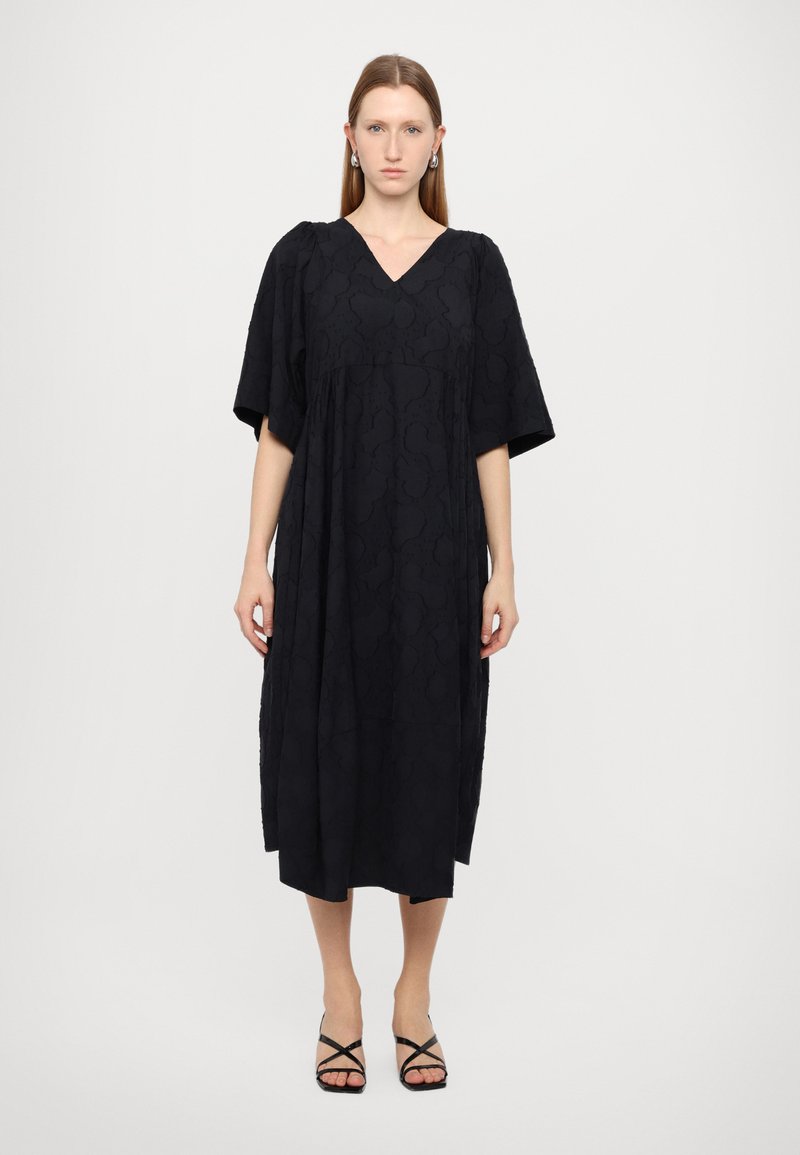 Zwart midi-jurk met een V-hals, korte wijde mouwen en een textuurstof. Gepaard met zwarte sandalen met bandjes.