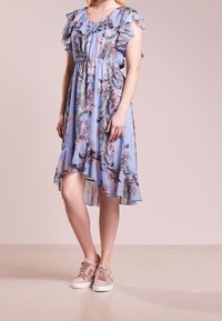 Vaaleansininen kukkakuosinen mekko, jossa on röyhelöhihat, joustava vyötärö, polvenpituinen helma ja läpikuultava päällikerros; yhdistetty vaaleanpunaisten lenkkareiden kanssa.