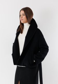 Giacca in misto lana nera con ampio colletto, design doppiopetto e tasche anteriori, indossata sopra un dolcevita bianco.