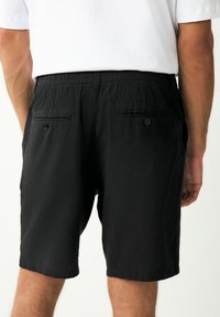 Pantalones cortos negros con una cintura elástica, bolsillos traseros con cierre de botón y una tela texturizada. El diseño es simple y casual.