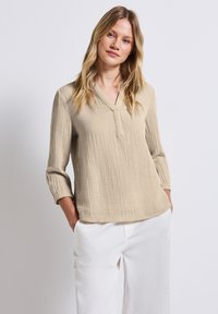 Beige, strukturierte Bluse mit 3/4-Ärmeln und V-Ausschnitt, dezenter Steppnaht, kombiniert mit weißen, locker sitzenden Hosen.