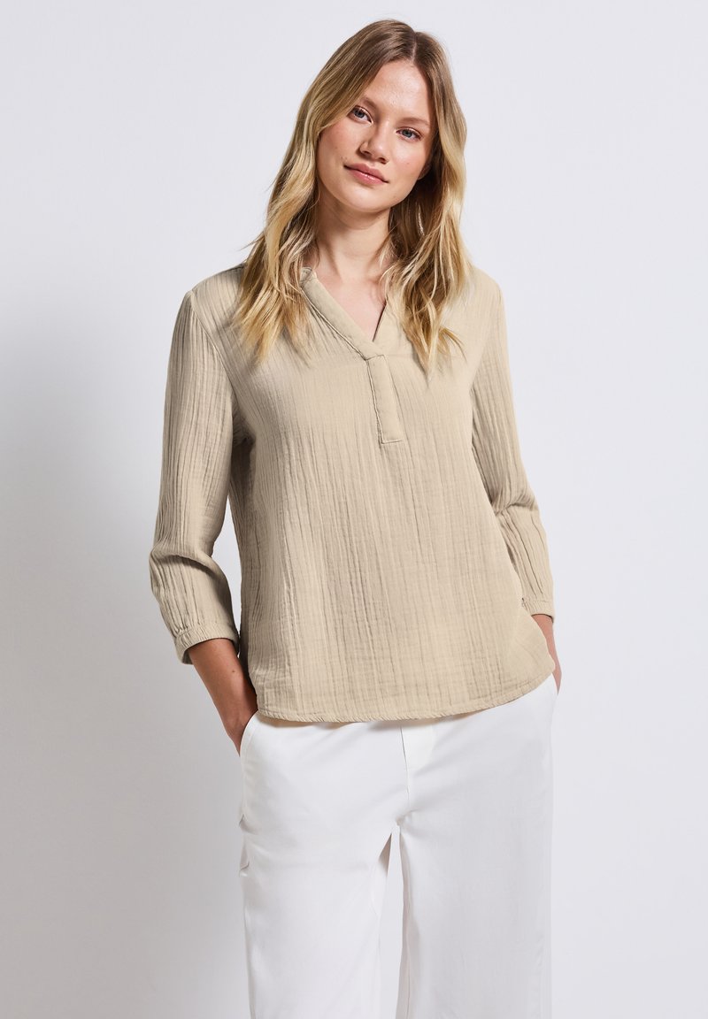 Beige, tekstureret bluse med 3/4 ærmer og v-udskæring, med diskrete stikninger, parret med hvide, løse bukser.
