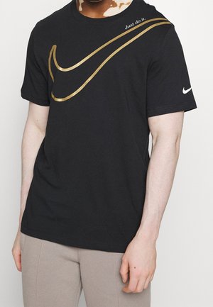 Osoba nosząca czarną koszulkę Nike z złotym logo Swoosh i napisem "Just do it." na piersi, w połączeniu z jasnobeżowymi spodniami.