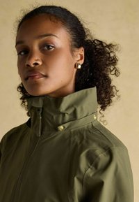 Jachetă impermeabilă de culoare verde măslin, cu guler înalt, având accente din metal auriu și închidere cu fermoar. Textură netedă, design minimalistic.