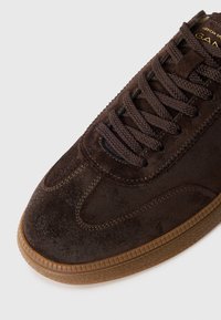 Brown suede sneaker with textured surface, round toe, and contrasting gum rubber outsole. Features matching brown laces and stitching accents.