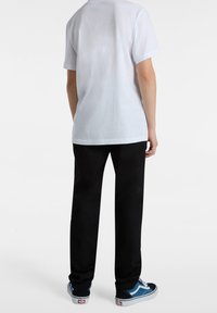 Vit bomullströja med korta ärmar, ihop med svarta byxor och blå-svarta sneakers, visar en enkel, avslappnad outfit bakifrån.