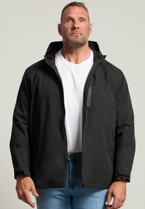 Homme aux cheveux courts et barbe, portant une veste noire zippée par-dessus une chemise blanche et un jean bleu, debout devant un fond clair uni.