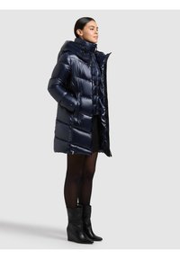 Tamsiai mėlynas pūkinis paltas su kapišonu, blizgiu, siuvinėtu tekstūros paviršiumi ir giliomis kišenėmis. Dėvimas prie juodų kulkšnies ilgio batų ir trumpos juodos suknelės.