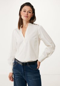 Witte blouse met lange mouwen, een plakkraag en ruches aan de randen. Draagtas met donkerblauwe spijkerbroek, voorzien van een zwarte riem. Glad textuur van de stof.