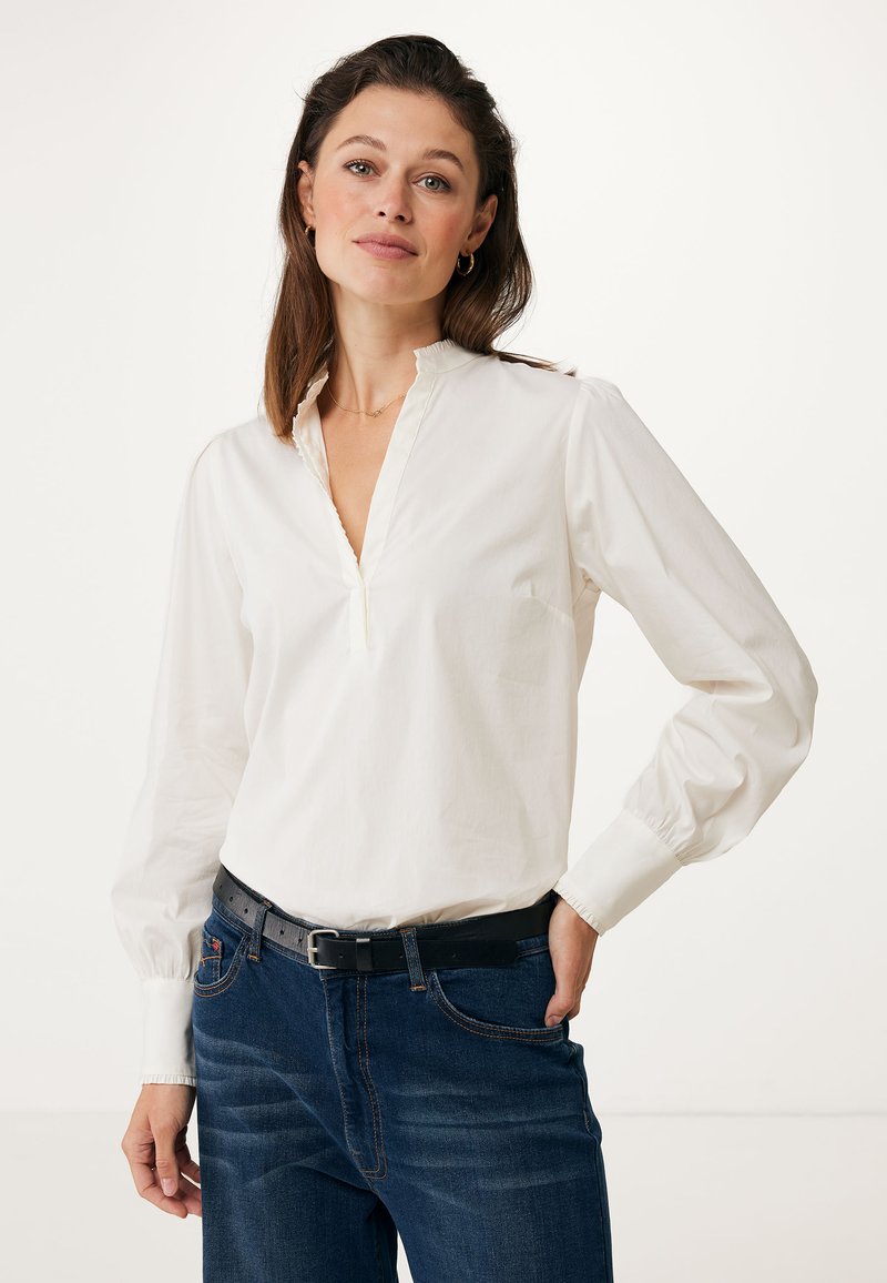 Witte blouse met lange mouwen, een plakkraag en ruches aan de randen. Draagtas met donkerblauwe spijkerbroek, voorzien van een zwarte riem. Glad textuur van de stof.