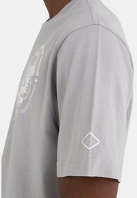 Hombre vistiendo una camiseta de manga corta gris claro con un gráfico blanco en el pecho y un pequeño logotipo de diamante en la manga, en un fondo liso.