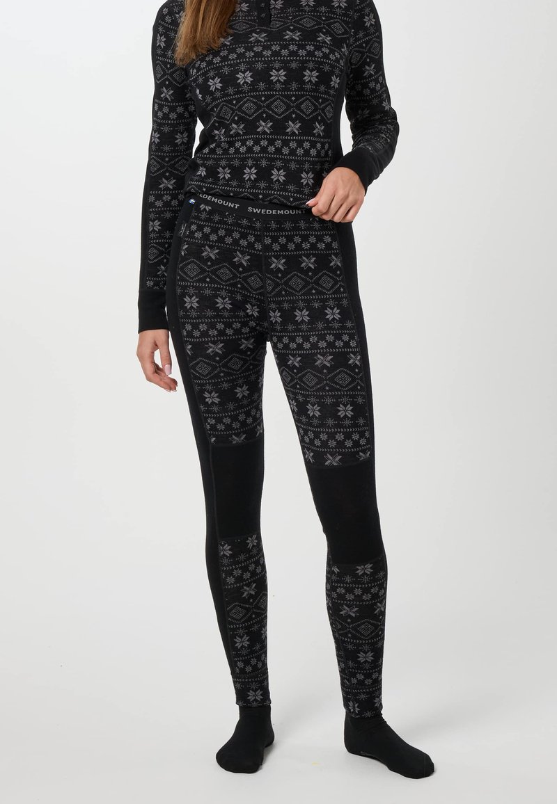 Personne portant un haut thermique noir à motifs et des leggings avec des dessins géométriques et des flocons de neige, debout devant un fond clair uni.