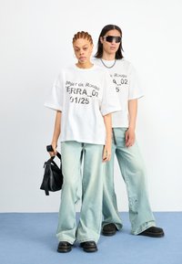 Two individuals wear oversized white t-shirts with black text and light blue wide-leg jeans. One holds a black handbag.