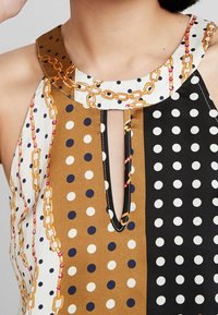 Top senza maniche multicolore con un motivo a pois e sezioni marroni, bianche e nere, e un design a catena al colletto.