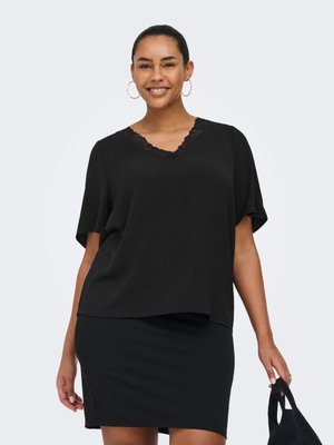 Femme souriante portant un haut noir à col en V bordé de dentelle et une jupe noire, tenant un sac à main noir sur un fond clair uni.