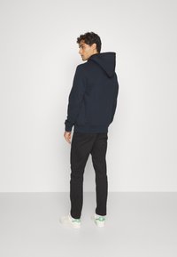 Felpa navy con tasca frontale, polsini e orlo a costine. Abbinata a pantaloni neri e sneakers bianche con dettagli verdi, vista da dietro.