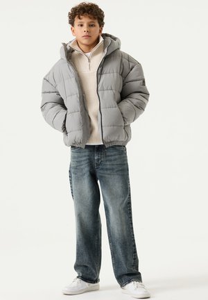 Junger Junge mit lockigem Haar steht frontal, trägt eine graue Steppjacke, einen cremefarbenen Zip-Pullover, weite blaue Jeans und weiße Sneaker.