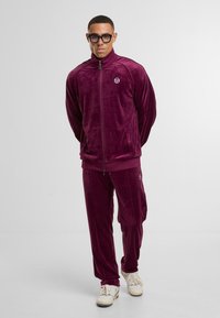 Mann in dunkelviolem Velours-Trainingsanzug mit Reißverschlussjacke und passender Hose, kombiniert mit weißen Turnschuhen und schwarzen Brillen, vor weißem Hintergrund stehend.