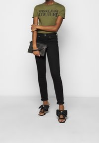 Olivově zelené tričko s potiskem "Versace Jeans Couture", černé skinny džíny, černá kabelka s cvočky a černé sandály s velkou mašlí.