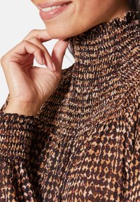 Braun gemusterter Rollkragenpullover mit geraffter Textur, versehen mit einem Rüschensaum und langen Ärmeln. Die Materialien wirken weich und leicht.