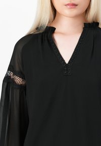 Blusa nera realizzata in tessuto leggero, con scollatura a V rifinita da pizzo e maniche trasparenti accentuate da dettagli in pizzo.