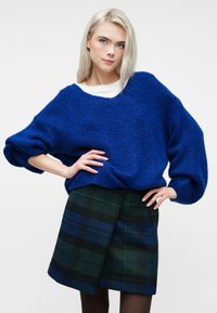 Kobieta z blond włosami w niebieskim, oversize'owym swetrze i zielono-niebieskiej spódnicy w kratę, stojąca z rękami na biodrach na białym tle.