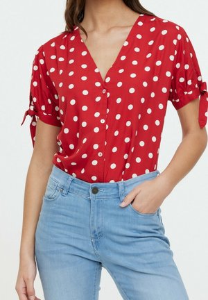 Nainen, jolla on punainen lyhythihainen pusero, jossa valkoisia pilkkuja ja hihoissa solmittavat yksityiskohdat, yhdistettynä vaaleansiniin korkeavyötäröisiin farkkuihin.