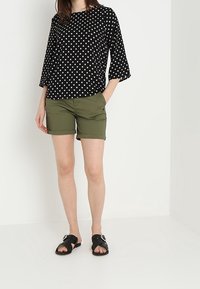 Kvinna i svart prickig topp, olivgröna shorts och svarta spännsandaler som står framför en vit vägg.