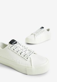 Sneakers bianchi in pelle liscia con suola in gomma texturizzata. Presentano lacci neri con testo stampato e un design a punta rotonda.
