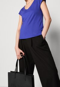 En person i en blå kortärmad topp och svarta byxor, som håller en svart Ted Baker tote-väska med ena handen i fickan.