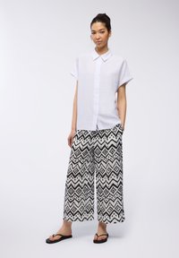 Camicia bianca a maniche corte con bottoni abbinata a pantaloni larghi bianco e nero con motivi geometrici. Il modello indossa ciabatte nere.