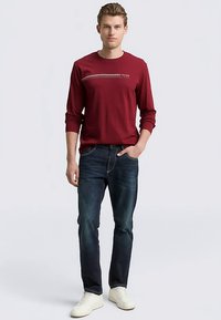 Maroon long-sleeve shirt with a contrasting horizontal white stripe across the chest, paired with dark blue jeans and white sneakers.