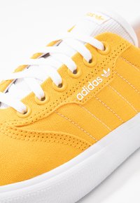 Gul kanvasfritidssko med vita snören, öglor och kontrasterande vit sula. På sidan finns tre vita ränder och logotypdetaljer.