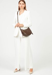 Borsa a spalla in pelle marrone con design curvo, tracolla regolabile e hardware minimalista, indossata con un completo bianco e décolleté beige.