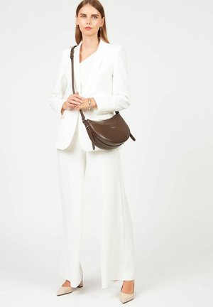 Borsa a spalla in pelle marrone con design curvo, tracolla regolabile e hardware minimalista, indossata con un completo bianco e décolleté beige.