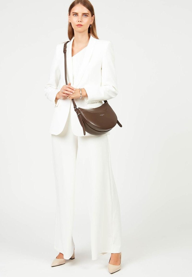 Borsa a spalla in pelle marrone con design curvo, tracolla regolabile e hardware minimalista, indossata con un completo bianco e décolleté beige.