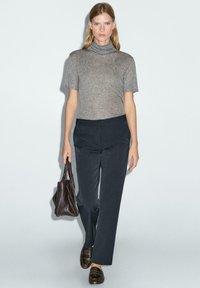 Grijze korte mouwen coltrui, donkere brede broek en bruine loafers. Bruine handtas in de hand, tegen een neutrale achtergrond.