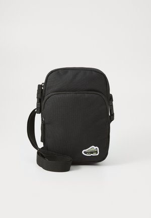 Kompakte schwarze Umhängetasche mit vorderer Reißverschlusstasche, verstellbarem Riemen und kleinem Sneaker-Patch in der unteren rechten Ecke.