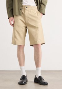 Beige bomullsshorts med sidelommer og rett snitt, kombinert med svarte loafers og hvite sokker. Minimalistisk design uten mønstre.
