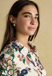 Young woman with long dark hair wears gold hoop earrings and a floral patterned button-up shirt, looking confidently to the side.