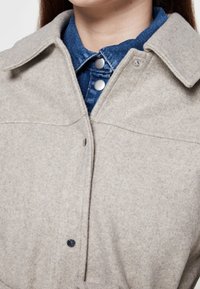 Chaqueta camisa de mezcla de lana de color gris claro con cuello, botones de presión y detalles de mezclilla. Presenta una textura suave y detalles de costuras visibles.