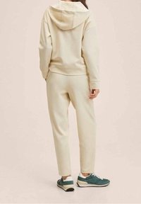 Cremfarbene Loungewear-Set mit Kapuzenpullover und tapered Pants. Weicher Stoff und entspannte Passform. Die Sneakers haben türkisfarbene und weiße Akzente.