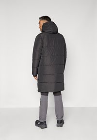 Manteau rembourré noir avec capuche, présentant un motif matelassé et une longueur plus longue. Porté avec un pantalon gris et des chaussures de sport noires.