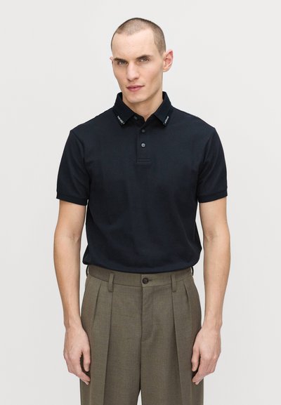 Kortärmad marinblå polo shirt med knäppning framtill och kontrasterande krage, tillsammans med högmidjade, ljusbruna byxor.